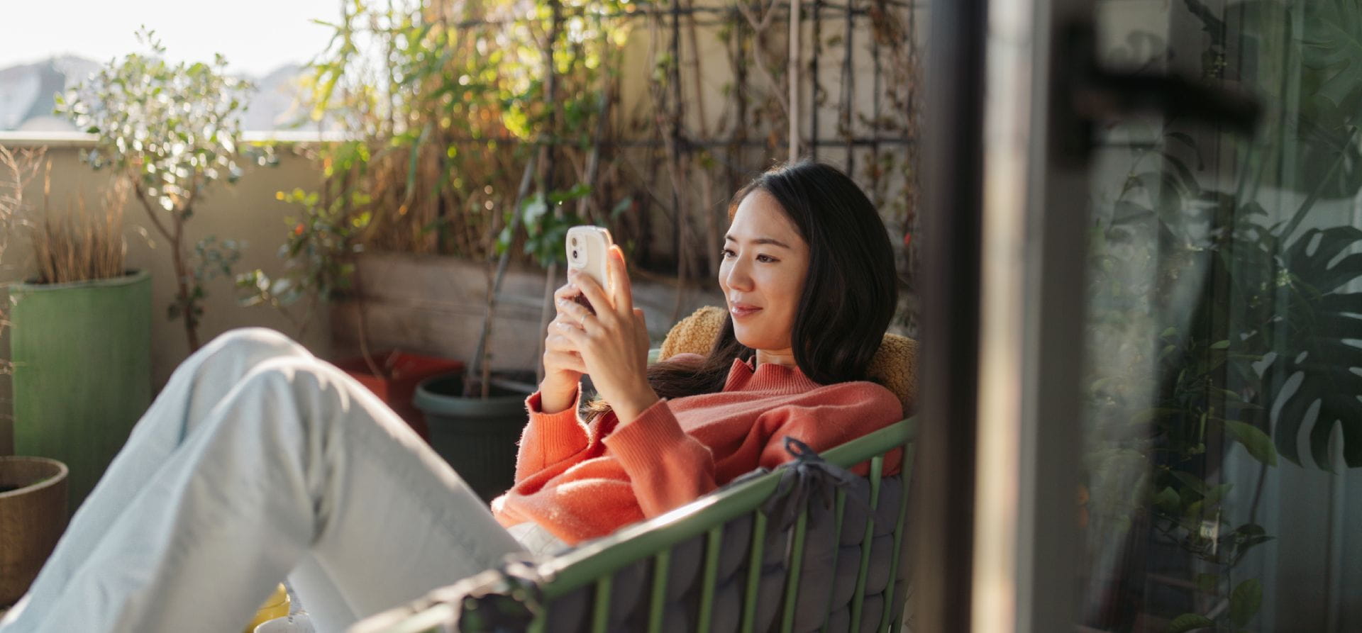 Eine Frau liegt in einem Sessel auf einem sonnigen Balkon und schaut lächelnd auf ihr Smartphone Eine Frau liegt in einem Sessel auf einem sonnigen Balkon und schaut lächelnd auf ihr Smartphone