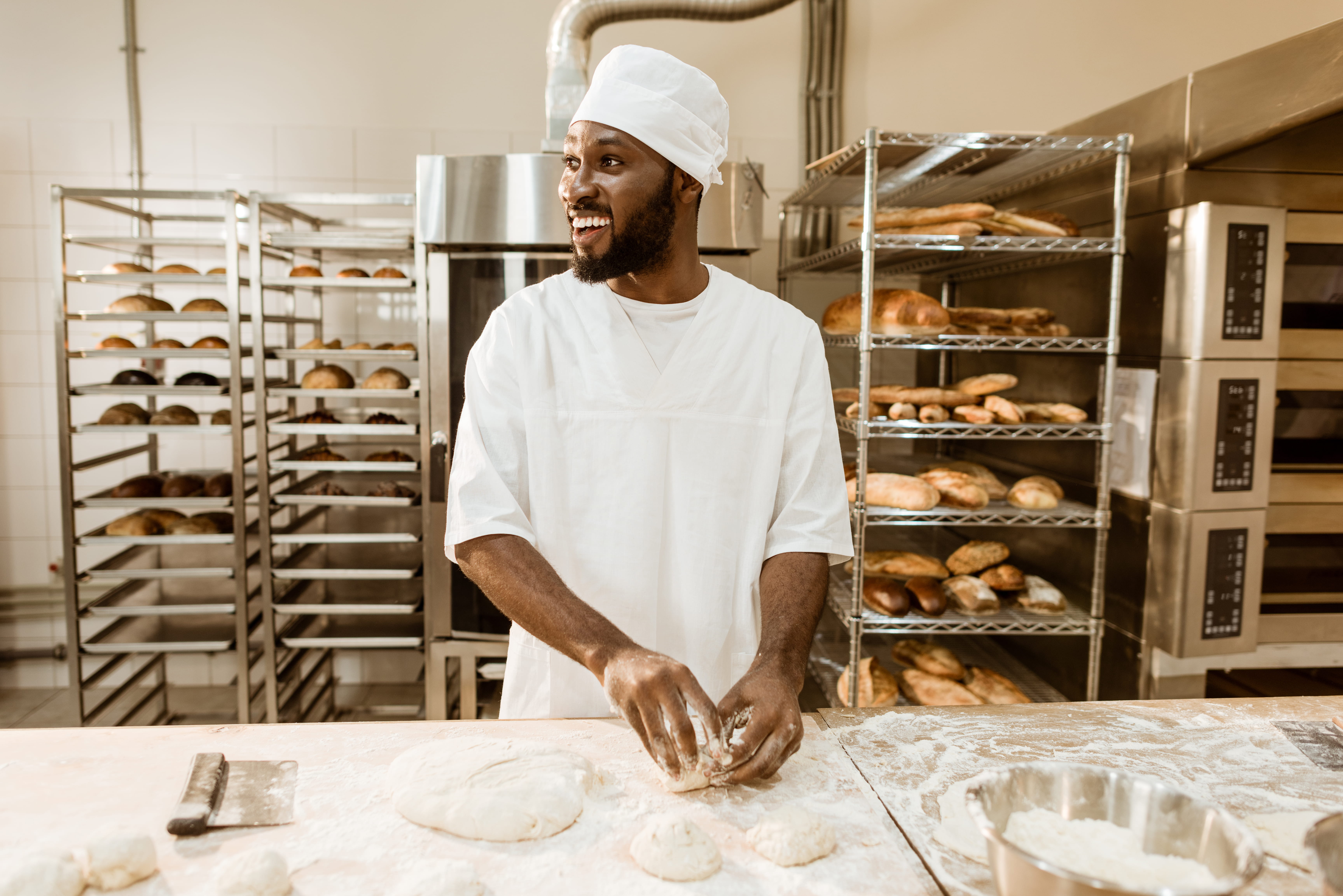 Baker in a white uniform and hat shaping dough on a floured surface in a bakery. Shelves behind are filled with various baked goods, including bread and pastries