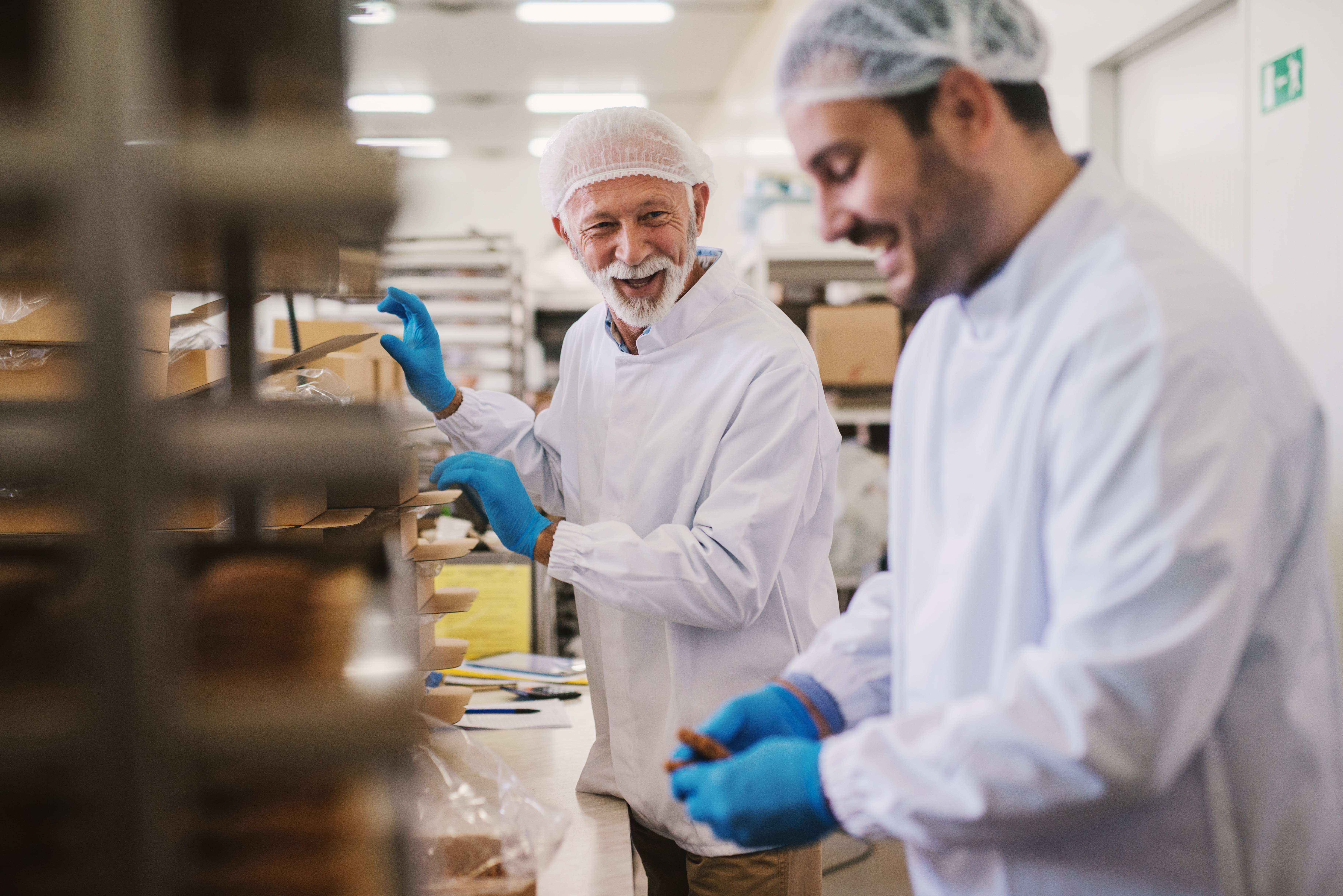 Two workers in white lab coats, hairnets, and gloves, smiling and working together in a bright, clean food production facility, handling packaged products