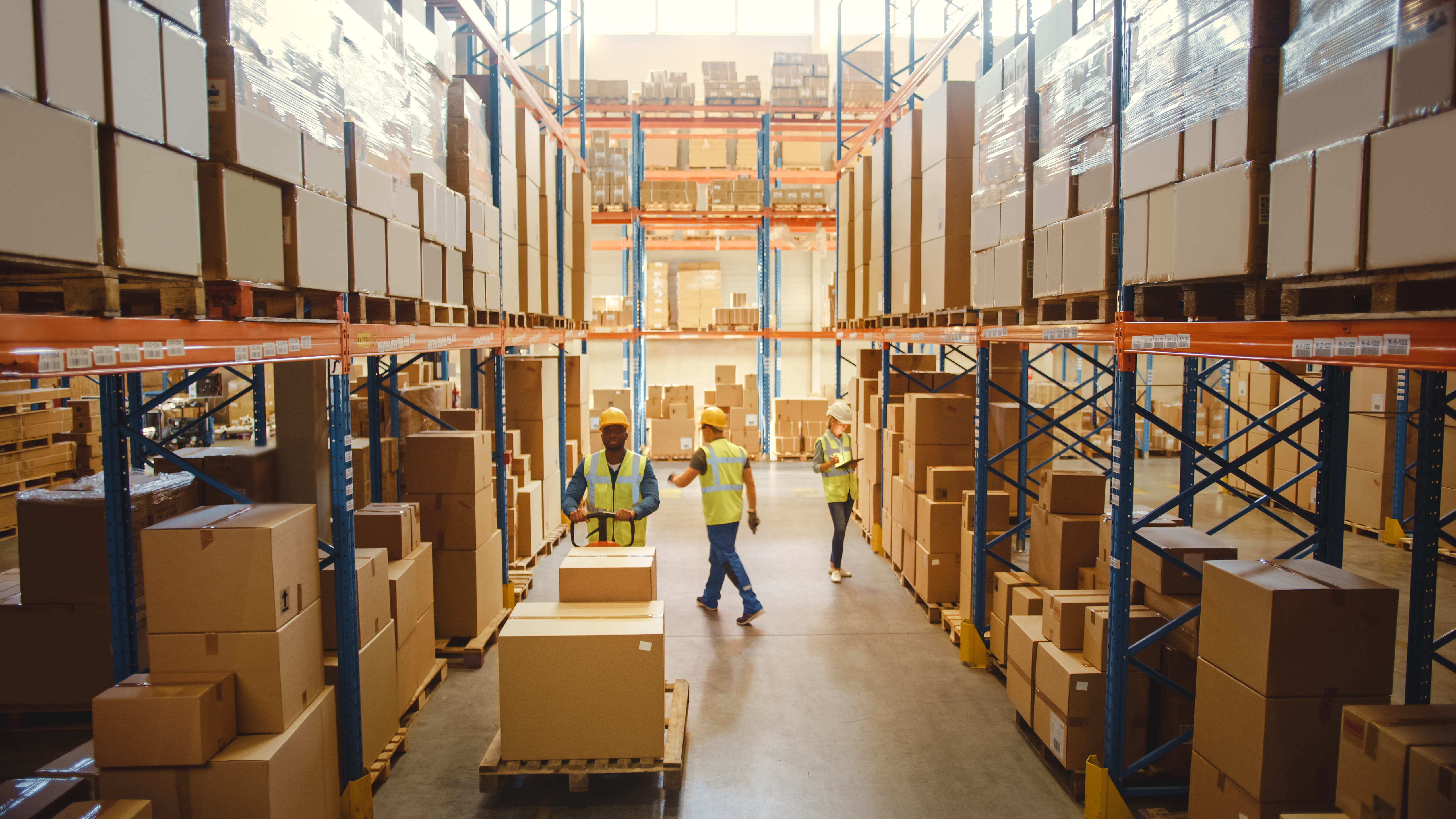 Large warehouse with high shelves filled with boxes and goods. Workers in high-visibility vests are organizing and moving boxes, ensuring efficient operations