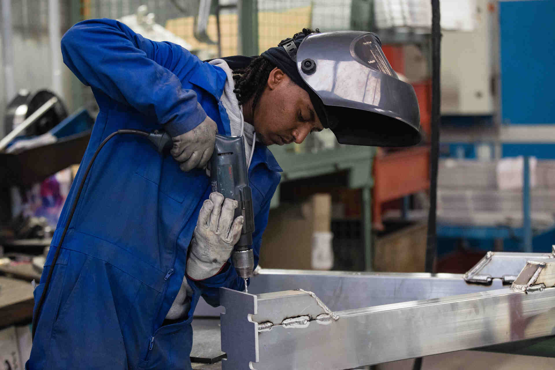 Worker in a blue coverall and protective helmet using a power tool to work on a metal piece in an industrial setting with machinery in the background.