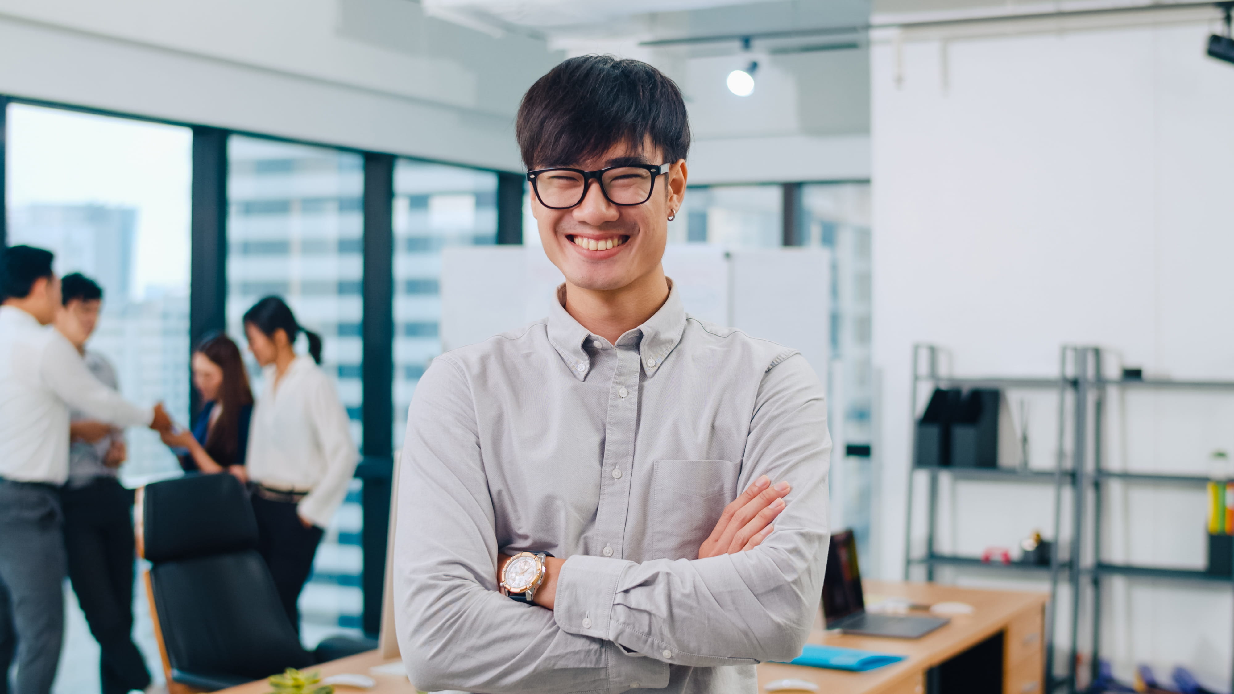 A smiling young professional with glasses and arms crossed standing in a modern office, with colleagues collaborating in the background.