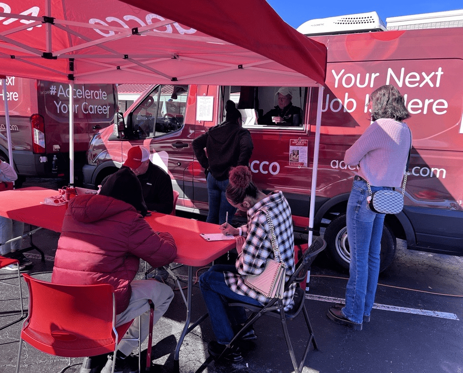 A group of job candidates sit at a table in front of two red Adecco Jobmobile vans.